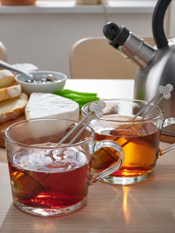 Na stole stoja dva sklené hrnčeky IKEA 365+ s čajom, za nimi je doska s nakrájanými syrmi.