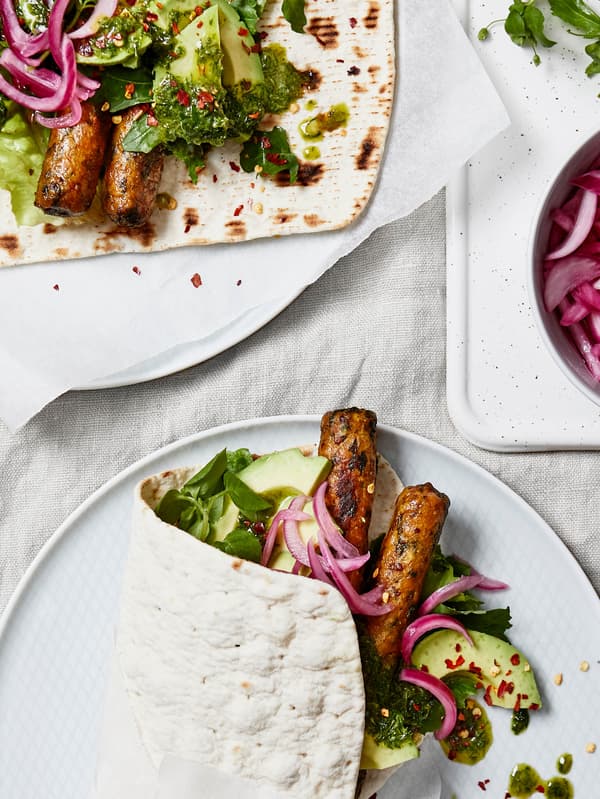 Na světlém ubruse leší dva bílé talíře s wrapy z tenkého chlaba GRÄDDAT, zeleninových hot dogů KORVMOJ, avokáda a červené cibule.