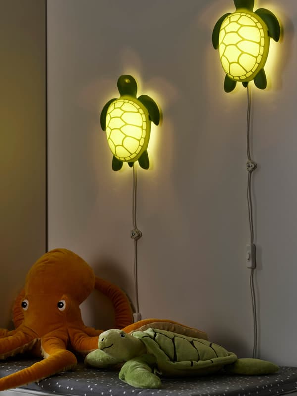 Na zdi svítí dvě LED lampy BLÅVINGAD ve tvaru želvy, pod nimi na komodě leží plyšové hračky, chobotnice a želva BLÅVINGAD.
