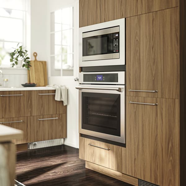 Na zdjęciu widać nowoczesną kuchnię z drewnianymi szafkami. W zabudowie znajdują się wbudowane mikrofalówka i piekarnik. Kuchnia jest czysta i zorganizowana, na blacie znajduje się deska do krojenia oraz kilka roślin. 