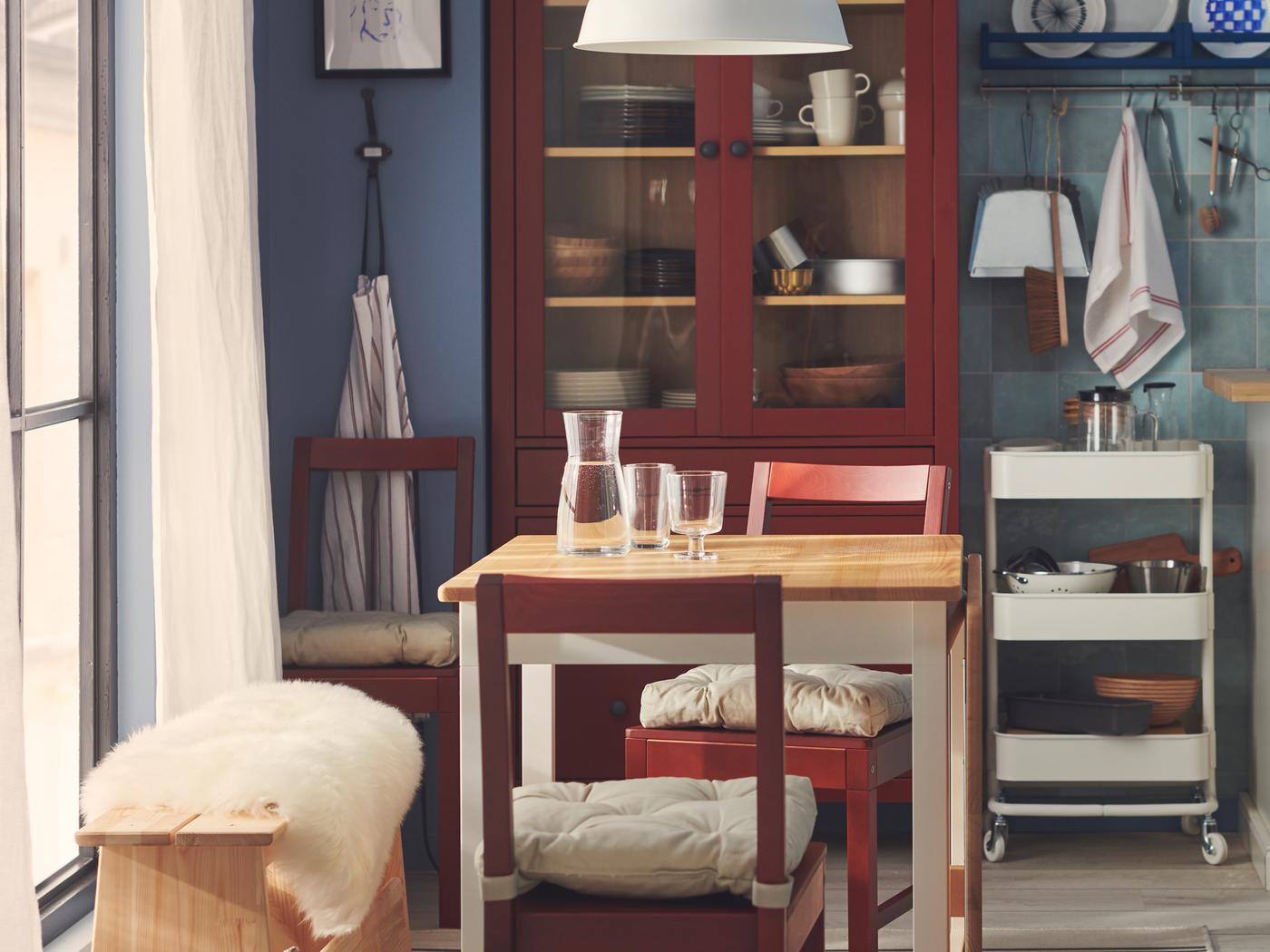 Naast een groot raam staan een tafeltje voor twee met witte poten en lichtbruin tafelblad en twee rode stoelen.