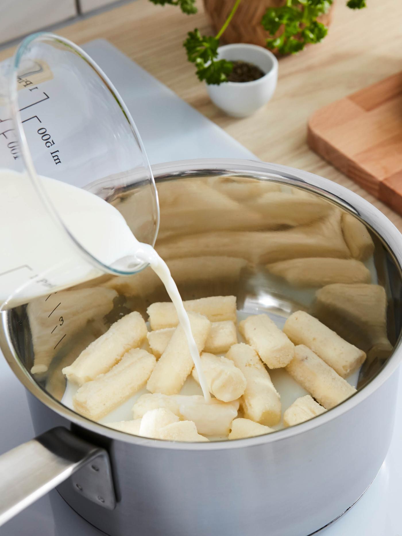 Načatý sáček masových kuliček HUVUDROL na prkénku ARTISTISK, vedle je pánev se smaženými kuličkami, v pozadí stojí miska brusinek.