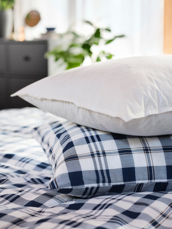 Nadýchaný vankúš LUNDTRAV položený na ďalšom vankúši a paplóne v károvaných posteľných obliečkach.