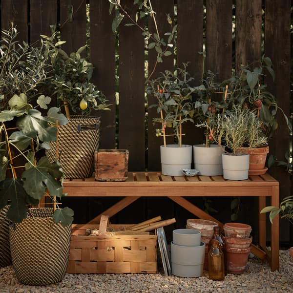 NÄMMARÖ outdoor bench at garden area in front of fence. Shows multiple plant pots being placed on top with plants and other items such as pots and baskets stored underneath. 