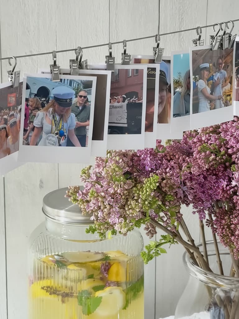 Närbild av polaroidbilder på en lina mot en vit trävägg. Bilderna visar ungdomar som firar, med studentmössor. I förgrunden en dryck med citron och syrenblommor.