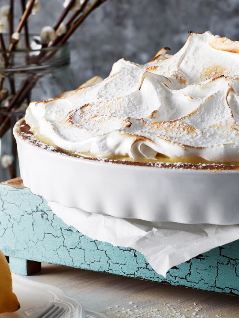 Närbild på citrontårta med maräng i en vit ugnsform. Står på ett påskdukat bord.