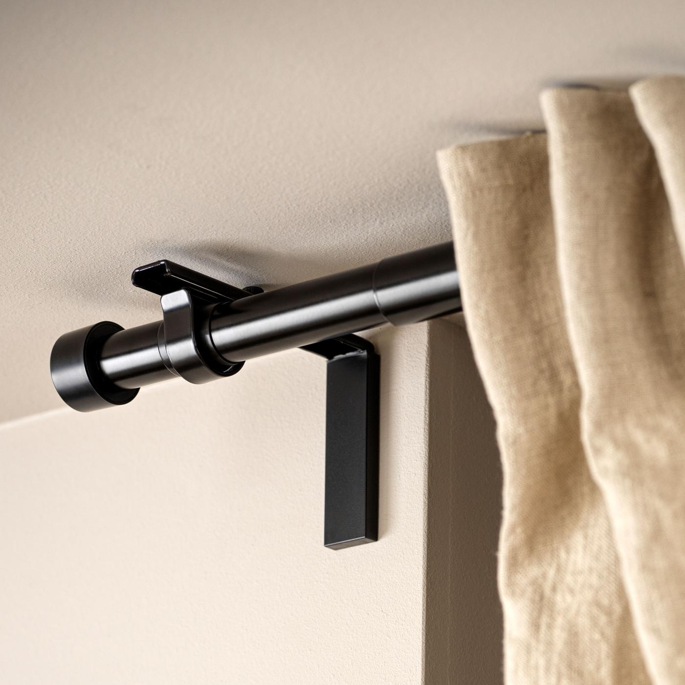 Närbild på en beige linnegardin, en svart gardinstång och dess väggfäste fäst på en beige vägg nära taket. 