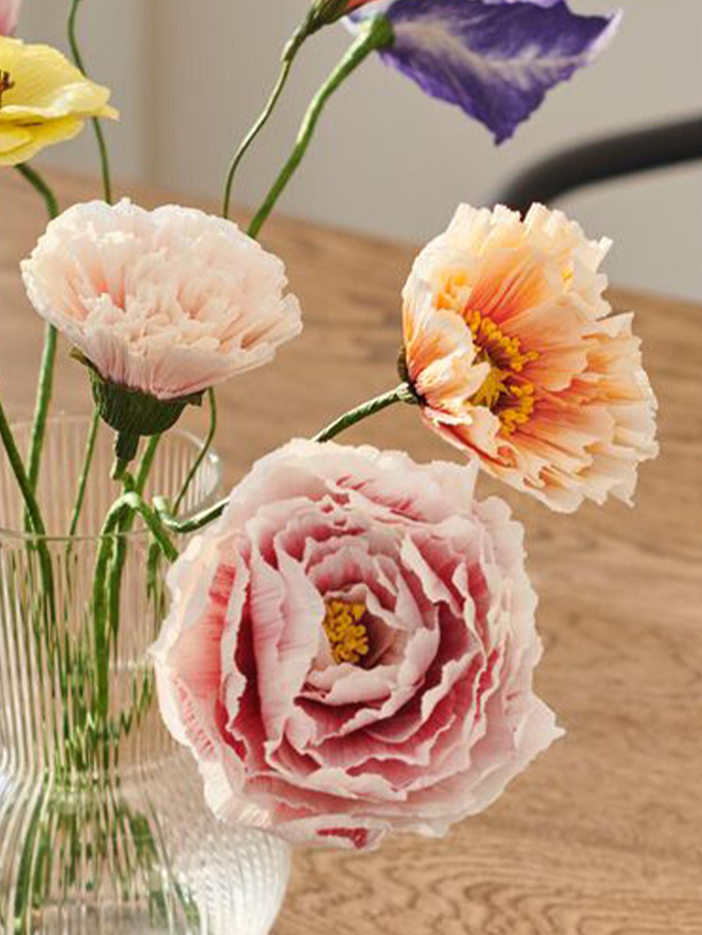 Närbild på en bukett med pastellfärgade pappersblommor i en glasvas på träbord. 
