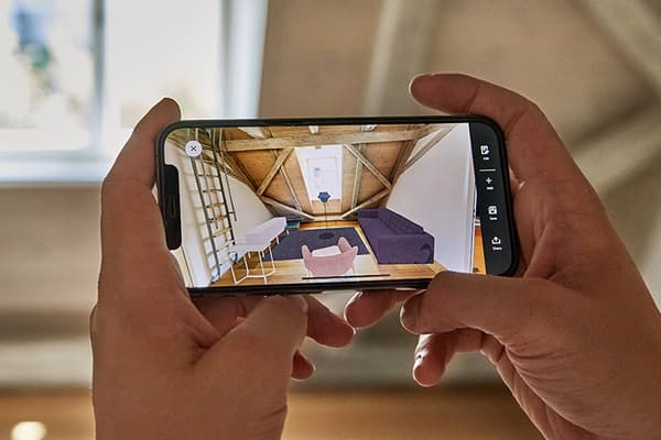Nærbillede af en persons hænder, der holder en smartphone, der er vippet, og som viser KREATIV-funktionen i IKEA-appen og visualiserer en møbleret stue.