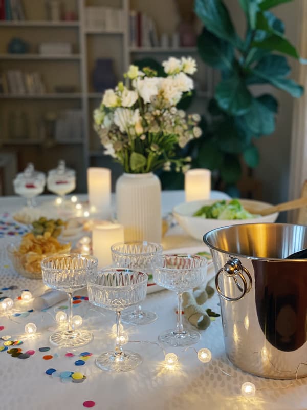 Nahaufnahme der Silvestertafel. Die Tischbeleuchtung bestehend aus einer Lichterkette und LED Kerzen reflektieren im Sektkühler. Buntes Konfetti ziert den Tisch. Kristallene Sektschalen stehen in der Tischmitte.