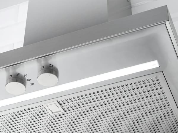 Nahaufnahme einer Dunstabzugshaube aus Edelstahl mit zwei Drehknöpfen auf der linken Seite, jeweils mit Zahlen und Symbolen beschriftet. Darunter befindet sich ein gelochter Metallfilter sowie ein rechteckiger Knopf in der Mitte des Panels.
