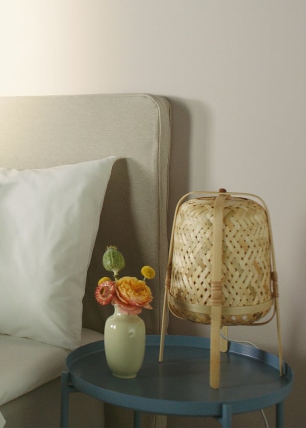 Nahaufnahme eines Bettes mit Kissen, eines Ablagetisches mit einer Leuchte und einer Vase mit orangefarbenen Blüten.