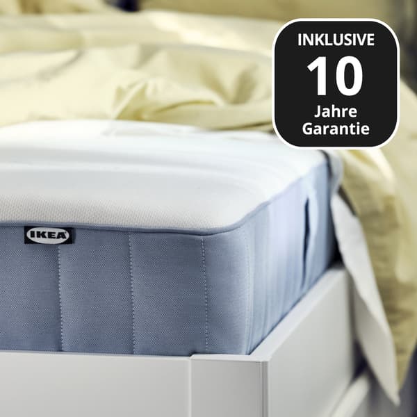 Nahaufnahme eines Betts mit Matratze und weißer Bettdecke. Die Matratze hat eine blaue Seitenfläche. Im Bild steht der Text ‚INKLUSIVE 10 Jahre Garantie‘.