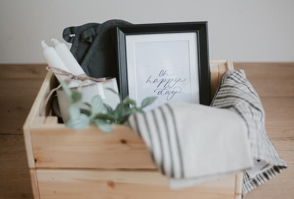 Nahaufnahme eines Geschenkkorbs bestehend aus einem KNAGGLIG Kasten aus massiver Kiefer bestückt mit Geschirrtüchern, Topflappen, weißen JUBLA Kerzen, einem shcwarzen Bilderrahmen und einer künstlichen Pflanze. 