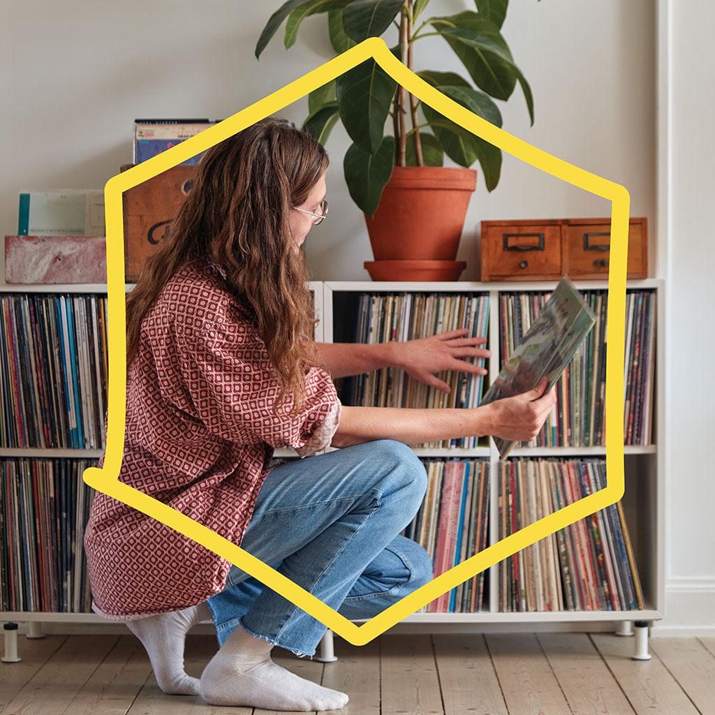 Nainen asettaa vinyylilevyä valkoiseen hyllyyn muiden levyjen joukkoon. Hyllyn päällä viherkasvi ja koriste-esineitä.