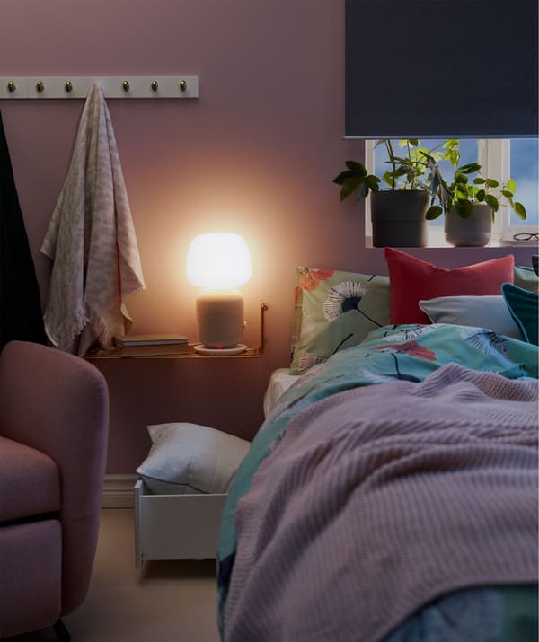 Narrow bedside shelf holding a SYMFONISK WiFi speaker table lamp glowing over a dim bedroom.