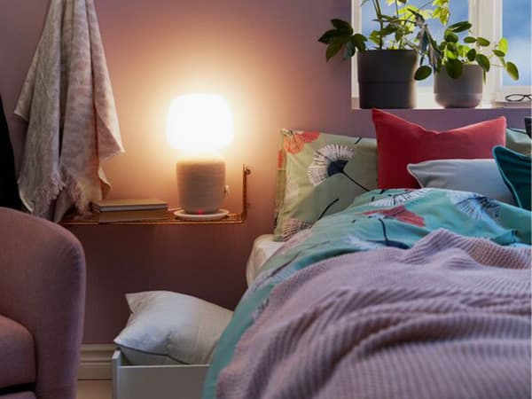 Narrow bedside shelf holding a WiFi speaker table lamp spreading a soft light over a dimly lit bedroom.