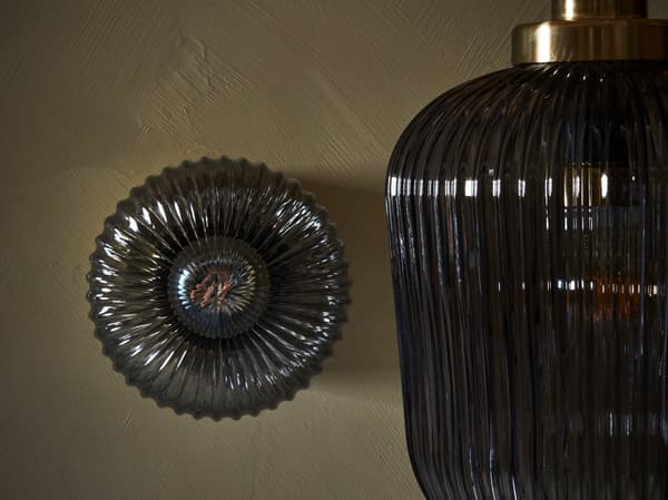 Nástěnná lampa a závěsná lampa SOLKLINT vyrobená ze šedého skla postavená u hnědé stěny.