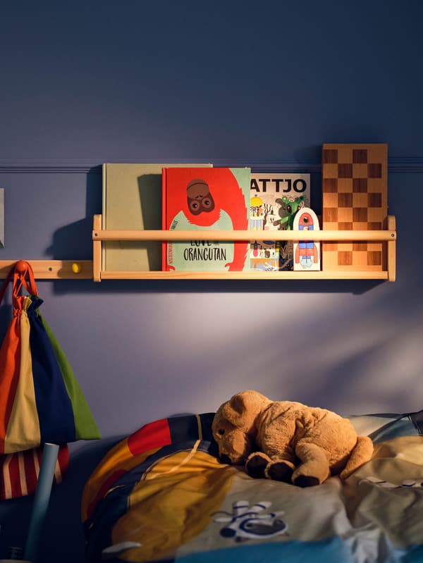 Nástenná polička FLISAT s knihami nad posteľou s plyšovým šteniatkom GOSIG GOLDEN.