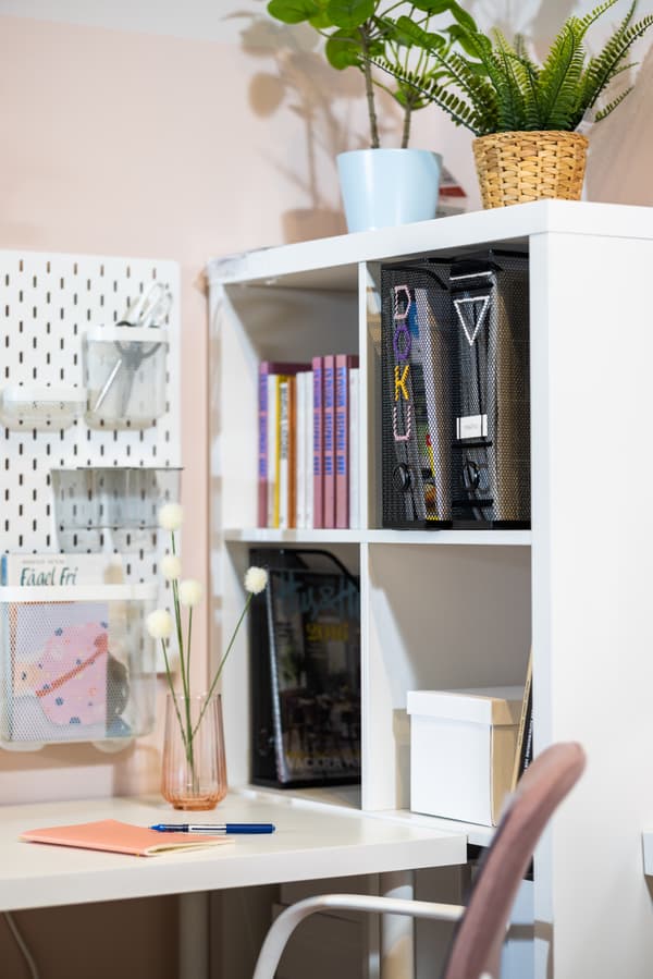 Neben einem Schreibtisch sthet ein Regal in dem schwarze Ordner und bunte Bücher stehen. Auf dem Regal stehen 2 bunte Vasen mit 2 grünen Pflanzen.