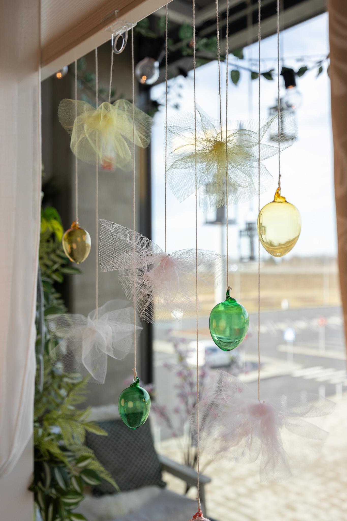 Небольшой круглый столик для детей, сервированный в пастельных тонах: тарелки, жёлтые салфетки, кружки и корзина с пасхальными яйцами; вокруг — ярко-жёлтые табуреты и декоративная зелень.