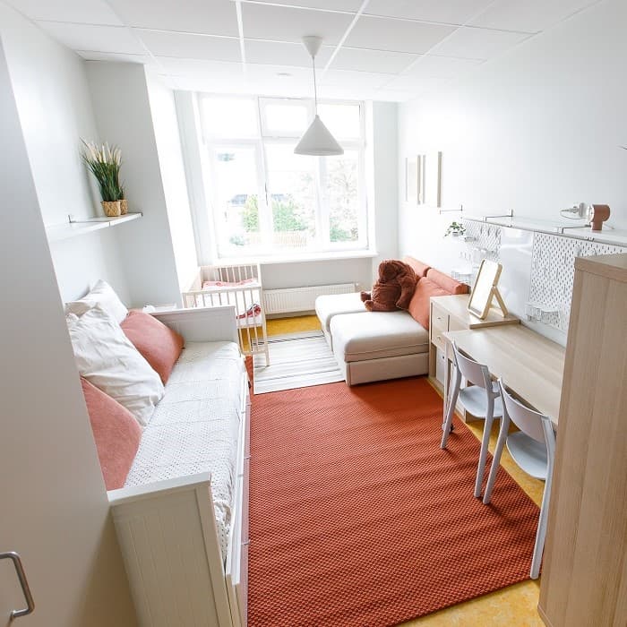 Nedidelis, tvarkingas kambarys krizių centre su viengule lova, oranžiniu kilimu, kompaktiška sofa, stalu ir kėde prie lango.