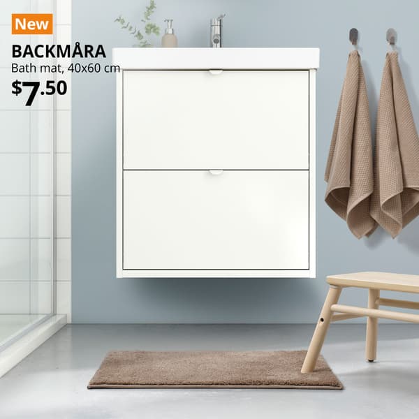 NEW tag. A beige BACKMÅRA bath mat is placed in the bathroom flooring right infront of the bathroom sink cabinet with a birch VILTO step stool placed on it. Black text on image reads "BACKMÅRA, Bath mat, 40x60 cm. Price: $7.50"