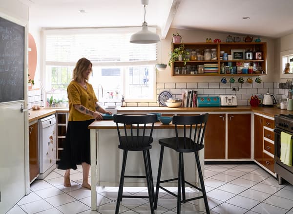 Nici ยืนอยู่ที่โต๊ะเตรียมอาหารซึ่งมีสตูลบาร์สีดำสองตัวในครัวที่มีตู้ครัวสีน้ำตาล ชั้นวาง และหน้าต่างบานใหญ่