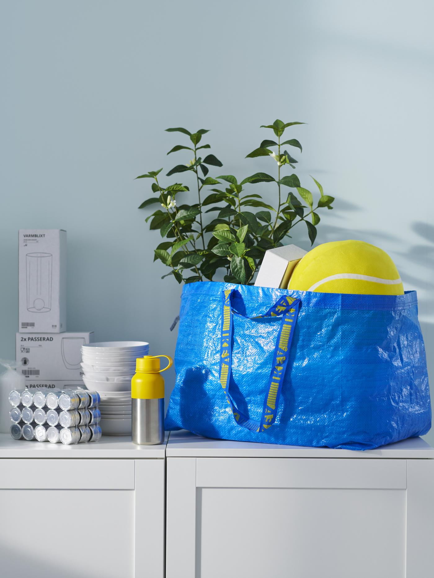Niebieska torba IKEA stoi na szarym regale. W środku są zakupy w tym piłka i roślina doniczkowa.