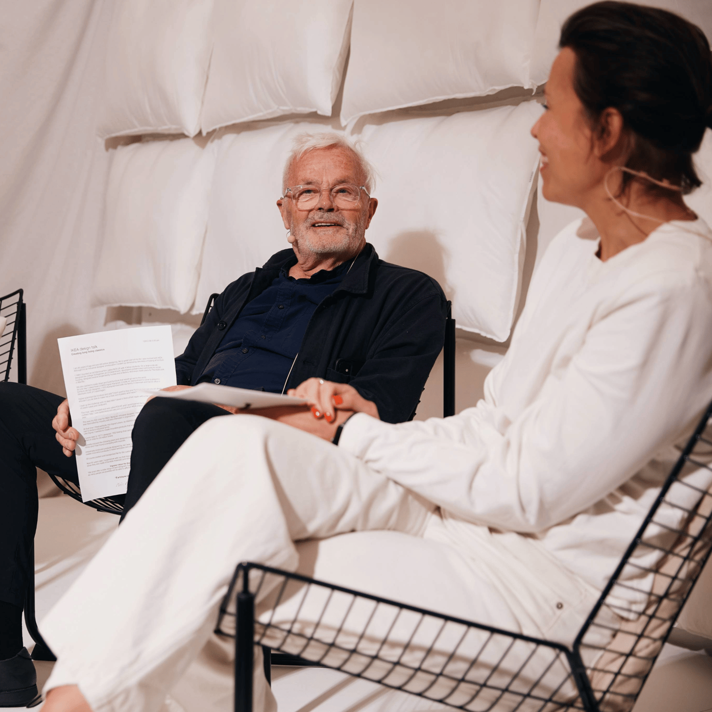 Niels Gammelgaard sidder og taler med en kvinde i sorte stole bag dem hænger hvide hovedpuder.