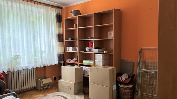 Nieuporządkowany, pomarańczowy pokój z regałem w imitacji drewna i stojącymi przed nim pudełkami kartonowymi.