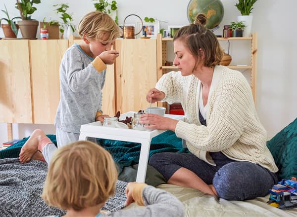 Nina et ses deux enfants en train de manger leur petit déjeuner au lit avec des armoires en bois à l’arrière-plan.