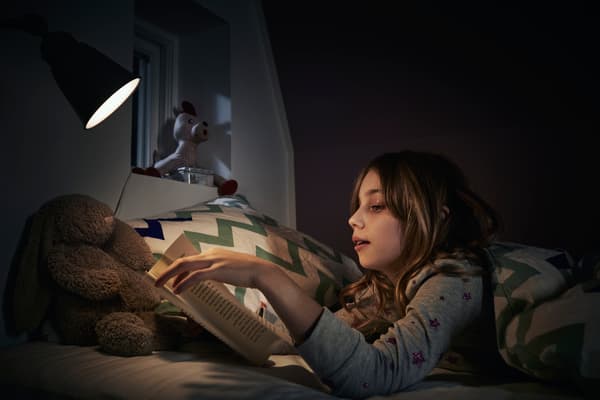 Niña leyendo libro con lámpara RYET apuntando a su cuento mientras está acostada en la cama.