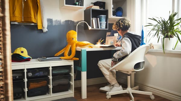 Niño estudiando en un escritorio en habitación infantil. Pulpo de peluche, planta, mueble organizador, lámpara, estanterías y ropa.