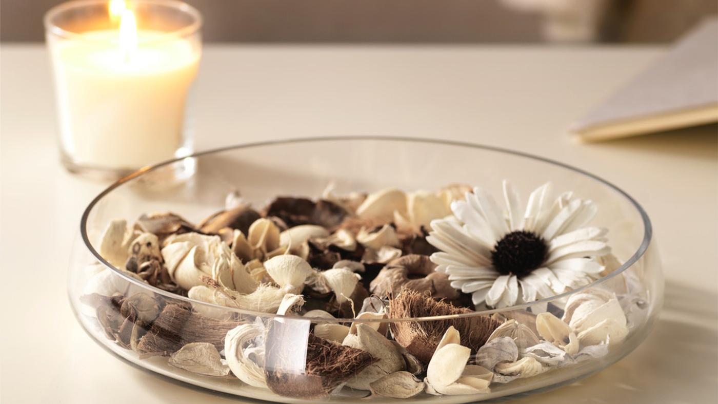 Nízka sklenená misa s potpourri vedľa ktorej je zapálená sviečka v skle.