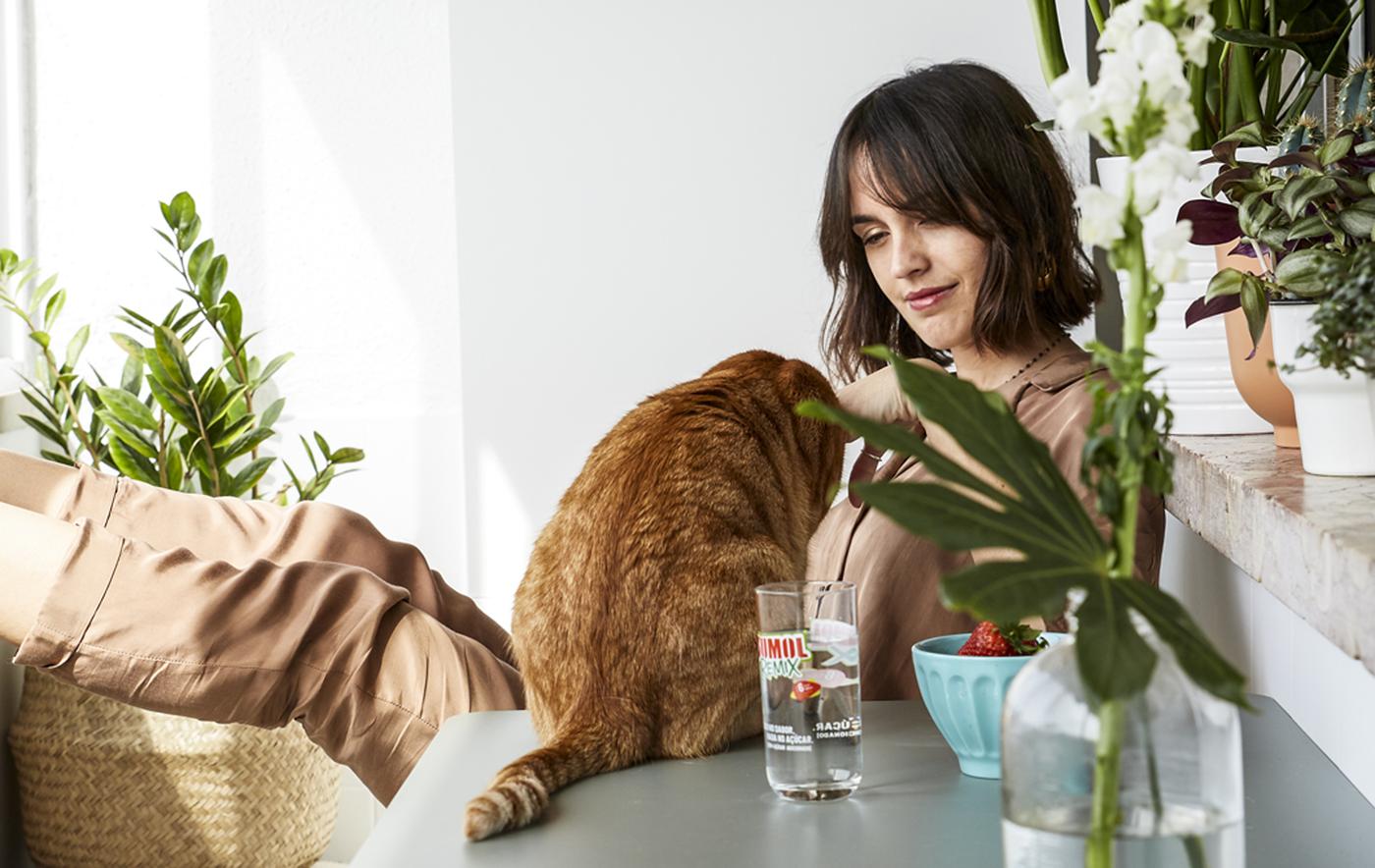 Nő és macska ül egy asztalnál egy fehér erkélyen, sok cserepes növénnyel.