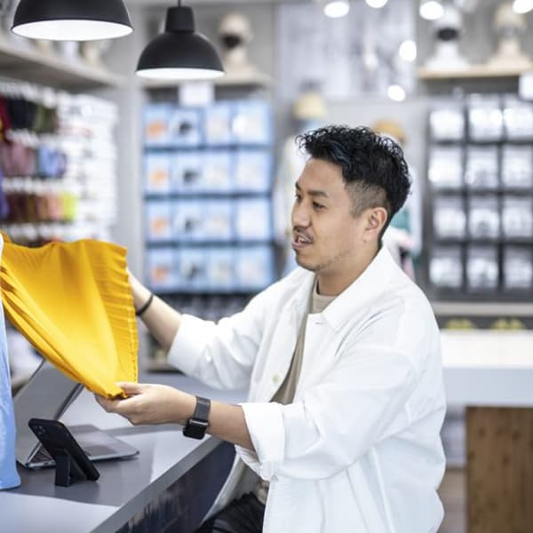 店舗のカウンターで人物が黄色い布を手に取り確認。背景には照明と棚に並ぶ商品があり、カウンターにはスマートフォンが置かれている。