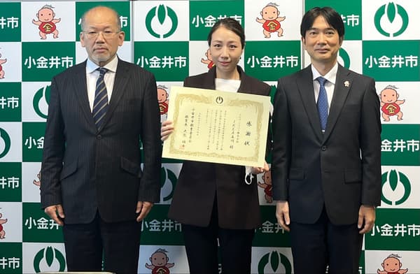小金井市のロゴ入り背景の前で、スーツ姿の3人が並び、中央の人物が賞状を持つ場面。