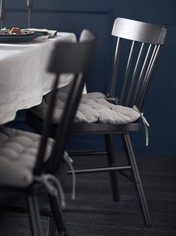 เก้าอี้ NORRARYD/นอร์ราริด สีดำพร้อมเบาะรองนั่งสีเทาตั้งอยู่ข้างโต๊ะที่จัดไว้พร้อมผ้าปูโต๊ะสีอ่อนในห้องที่มีผนังสีดำ