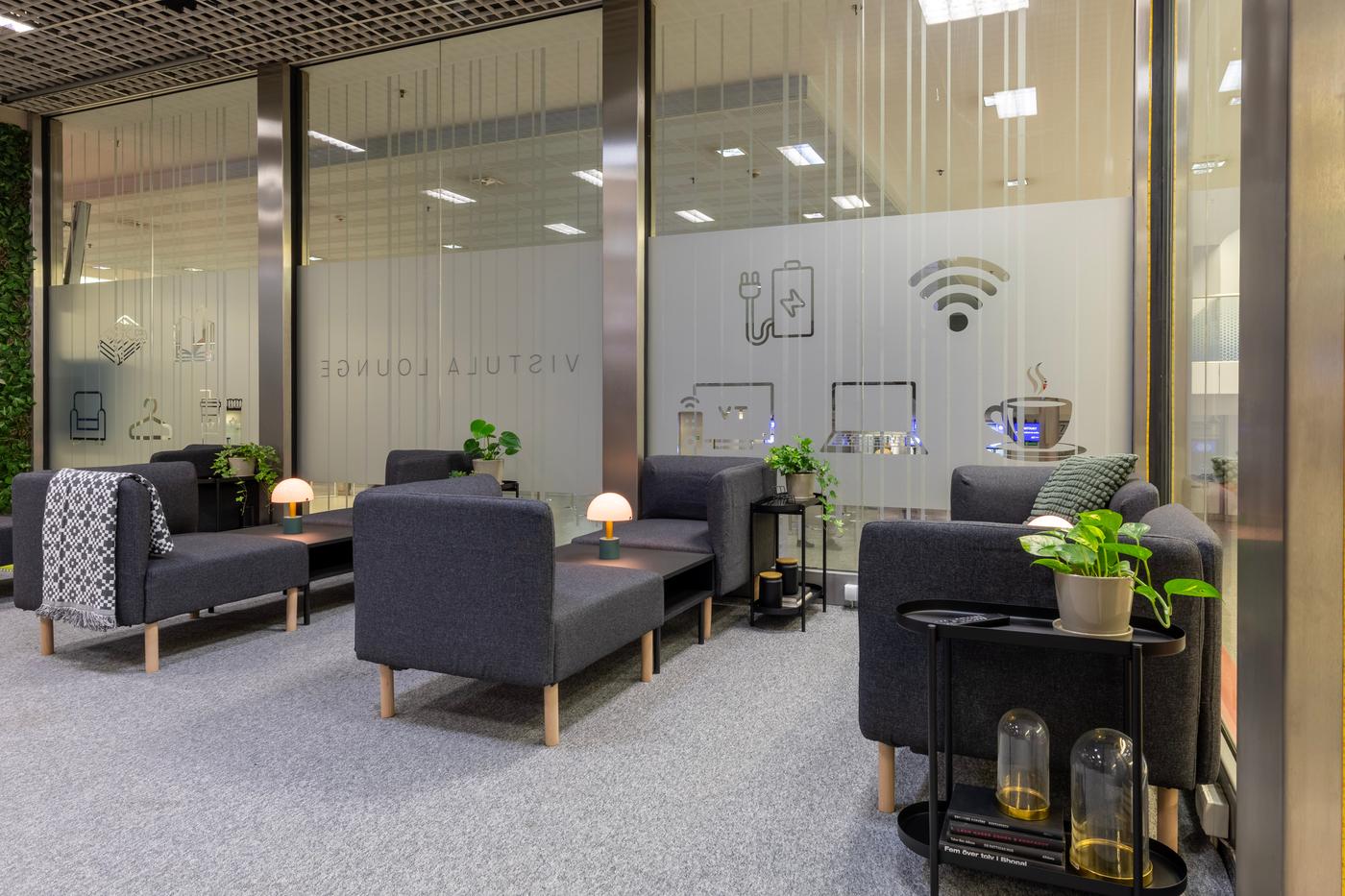 Nowoczesna poczekalnia z szarymi sofami, stolikami, roślinami i lampkami przy szklanej ścianie.