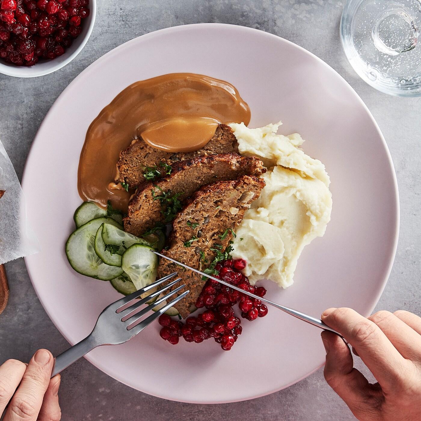 Nož i viljuška nad tanjirom s mesnom veknom, isečenim krastavcima, brusnicama, krem sosom i pire krompirom.