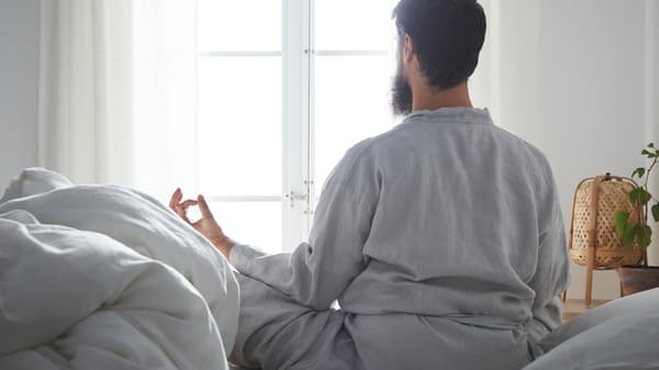 Num quarto de ambiente tranquilo, um homem com barba está sentado na cama de pernas cruzadas e em pose de meditação, a olhar pela janela.