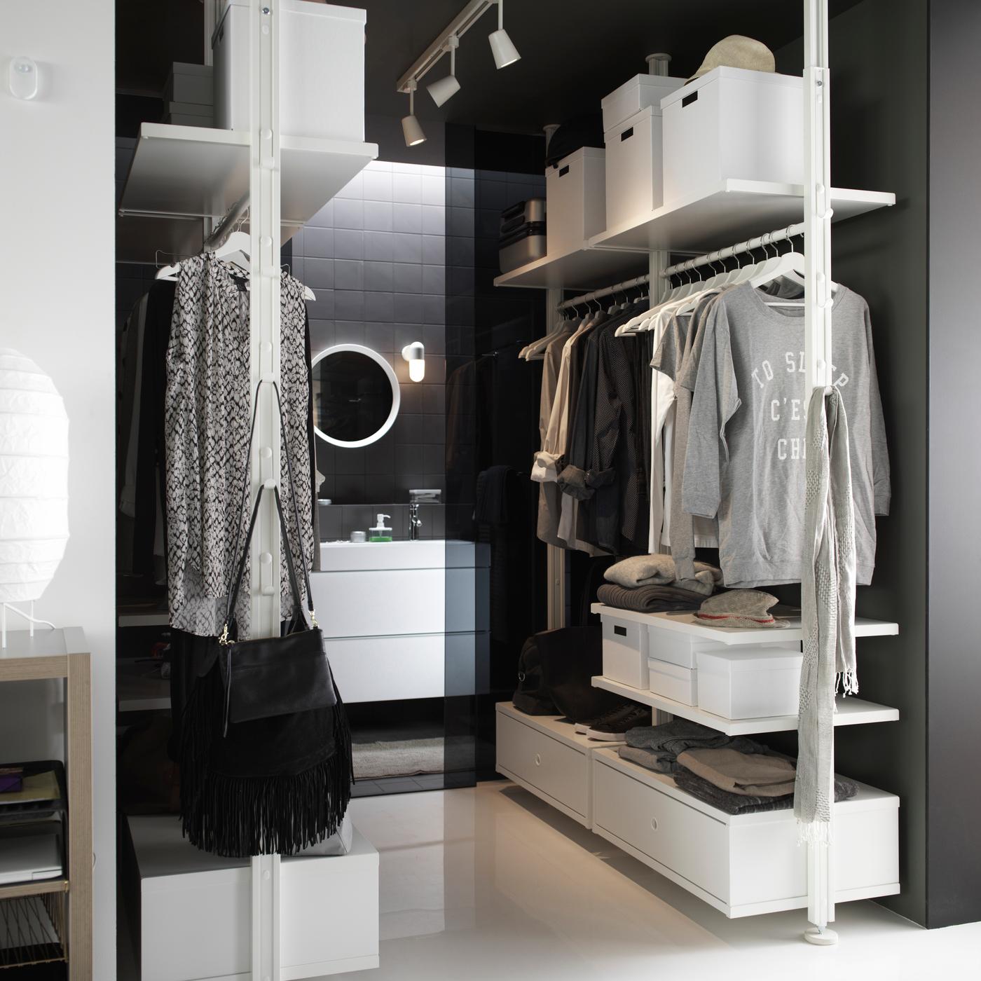 Nyitott gardróbrendszer modern belső térben fehér polcokkal, ruhafogassal, tárolódobozokkal és fiókokkal, a háttérben látható fürdőszoba kerek tükörrel.