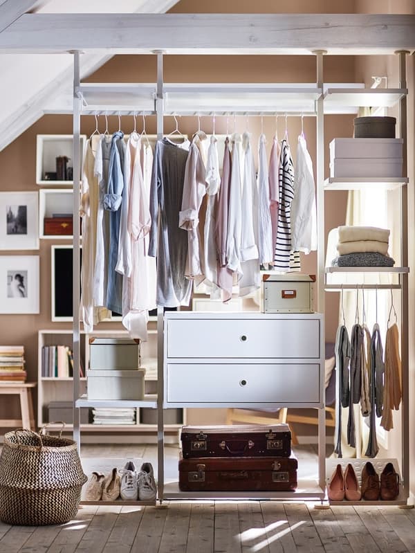 Nyitott ruhatári rendszer fehér polcokkal és fiókokkal, pasztellszínű ruhákkal teli akasztókkal, cipőkkel a padlón, tárolódobozokkal és egy ruháskosárral.