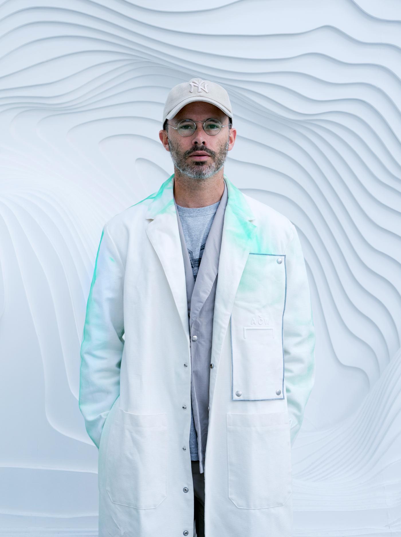 O artista Daniel Arsham, cunha bata de laboratorio con pintura turquesa, de pé sobre unha parede gris cunha textura de vagas.
