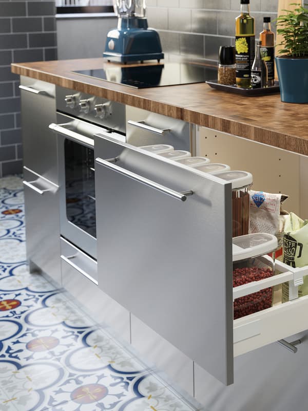 O bucătărie din inox cu un blat din lemn de stejar/furnir. Un sertar este deschis, în interior se află produse uscate.