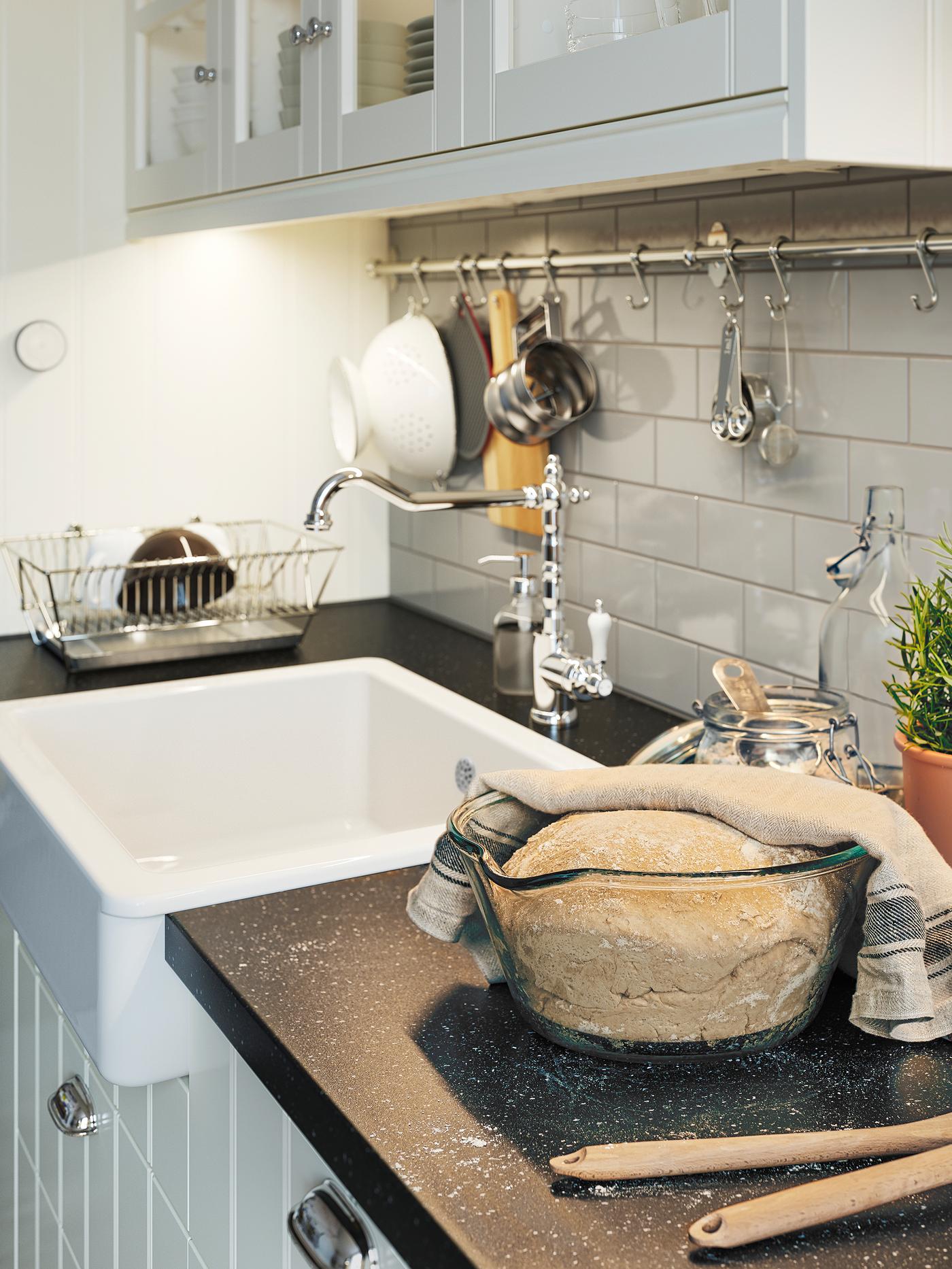 O bucătărie în nuanțe de alb în care un bol de sticlă cu aluat, acoperit pe jumătate, stă pe un blat negru cu efect mineral.