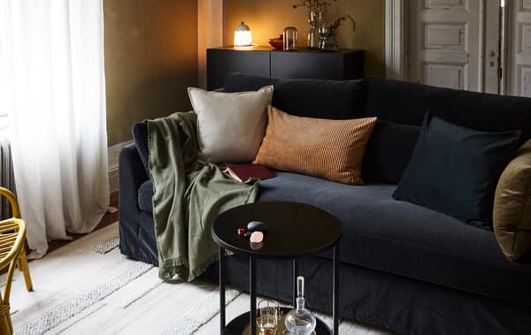 O cameră de zi cu o canapea, perne, pătură, covor, o măsuță, o comodă cu o veioză, vaze și un fotoliu din ratan.