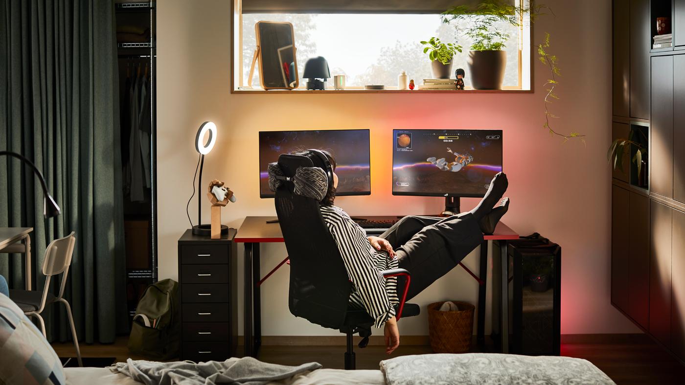 O cameră slab iluminată cu o persoană lăsată pe spate într-un scaun de gaming HUVUDSPELARE negru la un birou HUVUDSPELARE negru cu două monitoare.