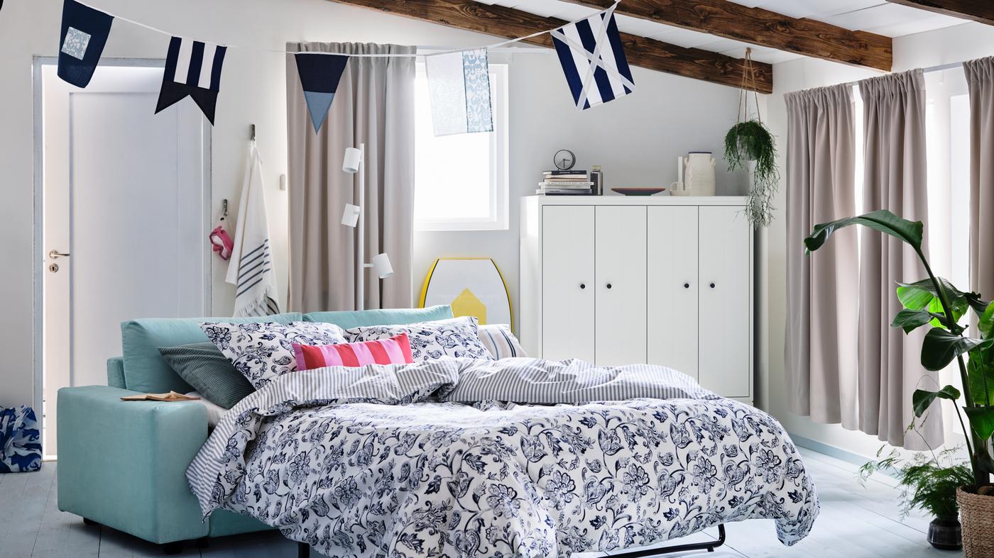 O casă de vară cu ferestre îmbrăcate cu draperii întunecate. O canapea extensibilă cu două locuri VIMLE, aranjată cu lenjerie de pat JUNIMAGNOLIA.